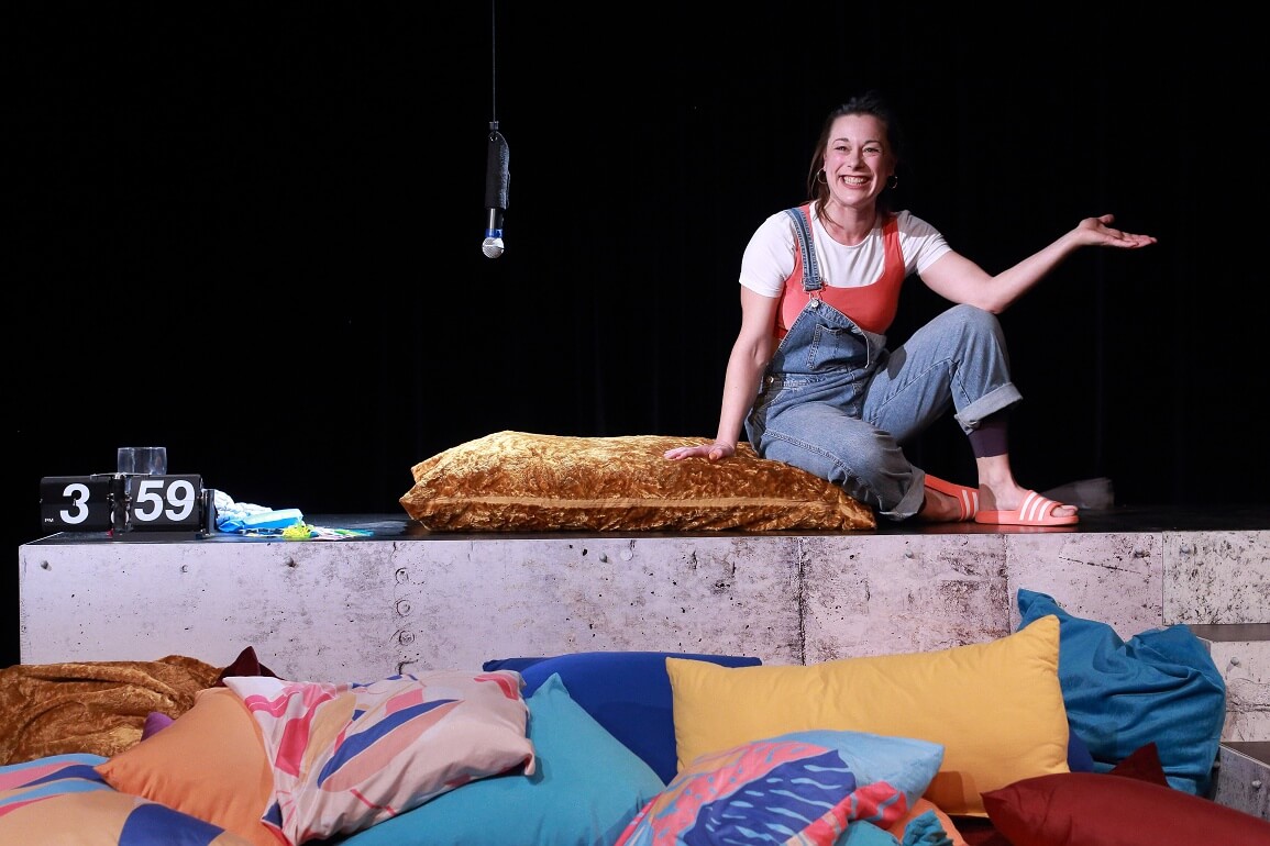 © Peter Schlipf Eine Frau sitzt auf einem Podest, vor ihr auf dem Boden befinden sich viele bunte Kissen. Auf dem Podest ist eine Uhr zu sehen und von der Decke hängt ein Mikrofon herab