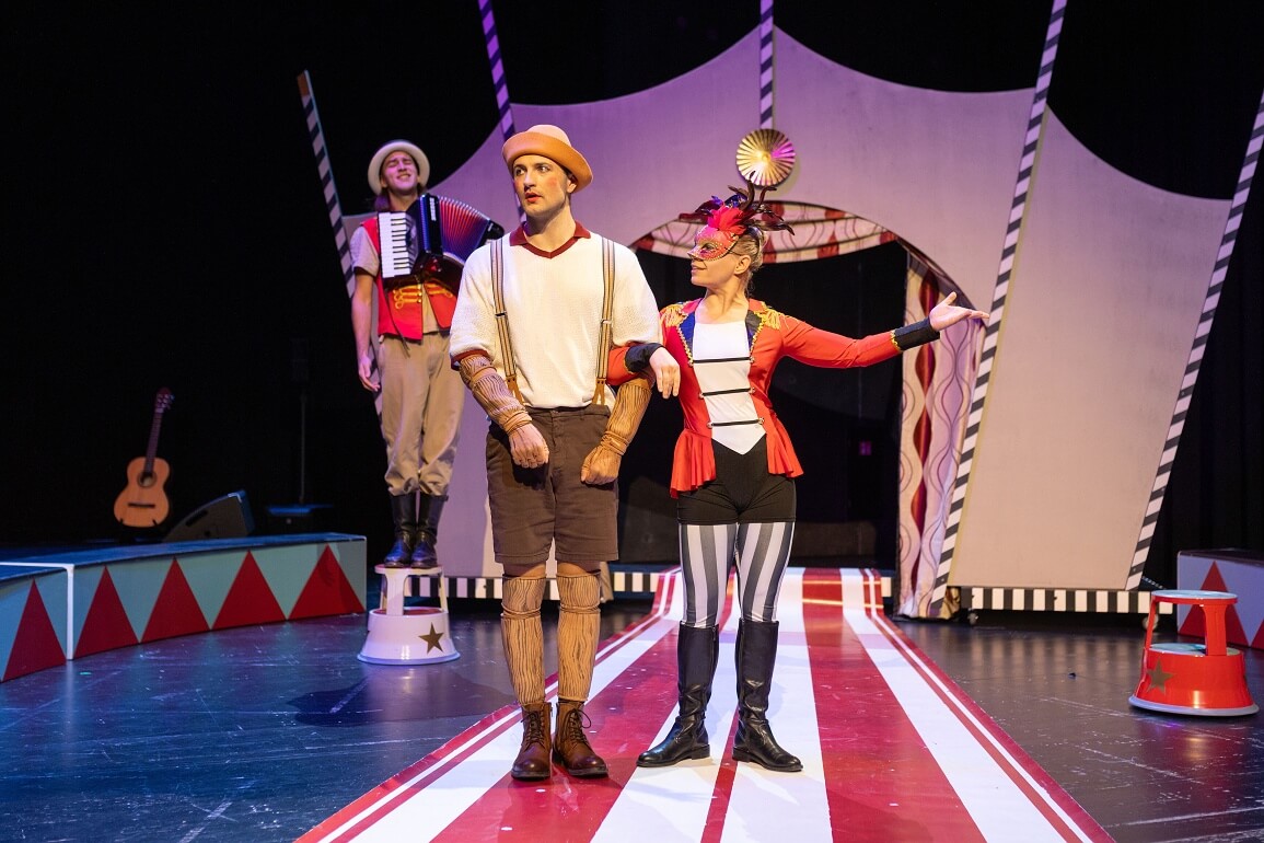 © Sofia Schomko Zwei Personen, eine in einem kurzen Hosenanzug, die andere in einem Artistenkostüm,stehen eingehakt vor einem Zirkus, im Hintergrund steht ein weiterer Zirkusartist mit einem Akkordeon und singt